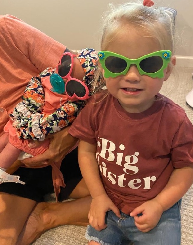 A little girl wearing sunglasses and a big sister t - shirt.